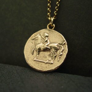 May include: A gold-toned circular pendant necklace featuring a textured surface with a relief design of a rider on horseback. The pendant is suspended from a matching gold-toned metal chain with rounded links, showcasing a detailed coin-like aesthetic.