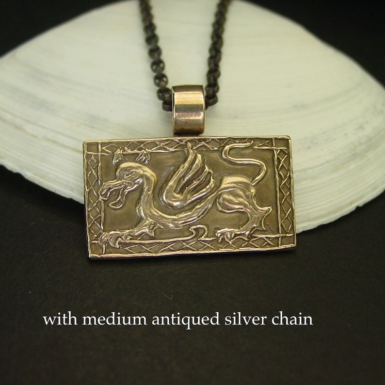 K&ouml;nnte beinhalten: Ein Bronzependant mit einem detaillierten gefl&uuml;gelten Drachen-Design, das in einem rechteckigen Rahmen mit einem keltischen Knotenmuster eingefasst ist. Der Anh&auml;nger ist an einer silbernen Kette.