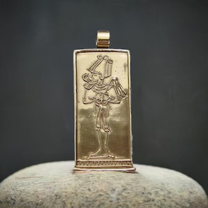 May include: Gold-colored rectangular pendant with an engraved Egyptian figure. The pendant features a detailed depiction of a pharaoh or deity, with intricate headdress and clothing. The pendant is suspended from a small loop at the top.