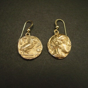 May include: A pair of gold-colored earrings. Each earring features a circular pendant with a detailed relief design. One pendant depicts an owl, and the other shows a profile of a person. The earrings have a simple hook design.