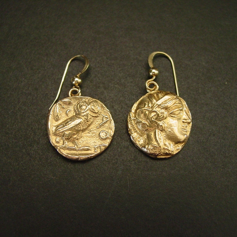 May include: A pair of gold-colored earrings. Each earring features a circular pendant with a detailed relief design. One pendant depicts an owl, and the other shows a profile of a person. The earrings have a simple hook design.