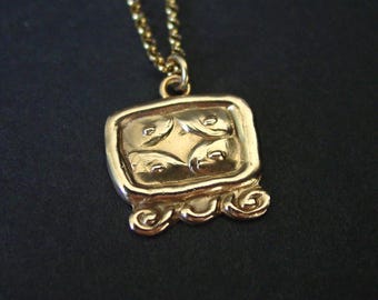 May include: A gold-toned pendant necklace featuring a rectangular charm with rounded corners. The charm displays a raised, symmetrical four-part embossed pattern and three decorative scroll-like elements along the bottom edge.