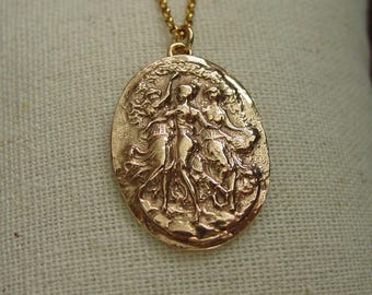 May include: A gold-toned oval pendant necklace featuring a raised relief design of three figures dancing in flowing garments. The pendant is suspended from a delicate gold-toned chain, showcasing intricate textures and a metallic finish throughout.