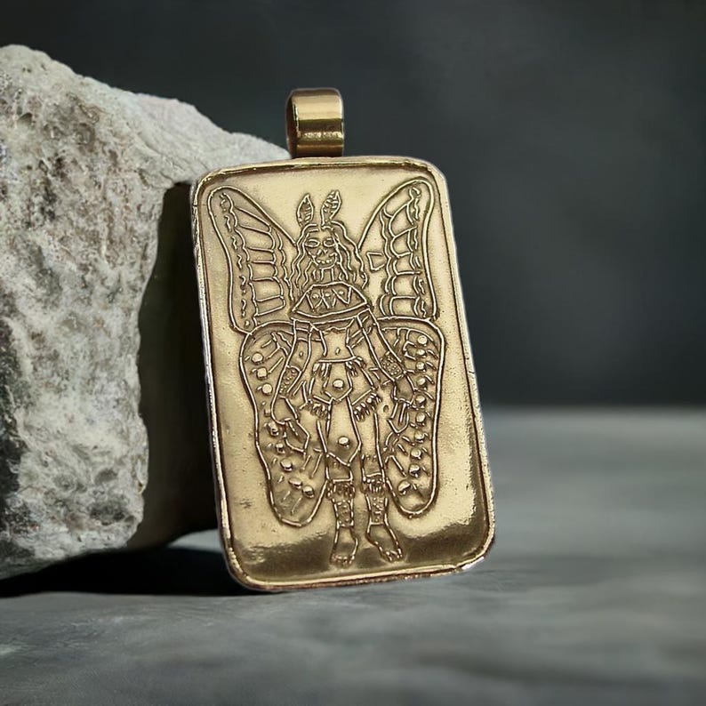Gold rectangle shaped pendant with tube bail on top. In its center is a female wearing traditional warrior Aztec outfit. She is holding weapons, had butterfly wings, has moth antenna on top of her head and her face is a skeleton.