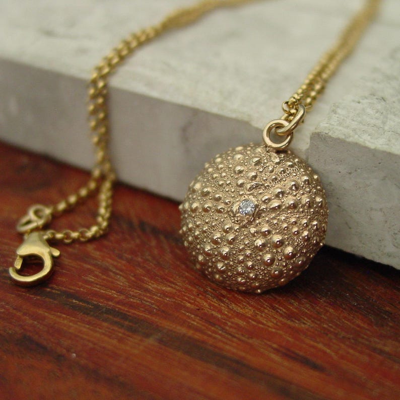 May include: A gold pendant necklace with a textured, spherical charm and a small gemstone. The charm is suspended from a fine gold chain with a lobster clasp. The necklace is presented on a light-coloured surface.