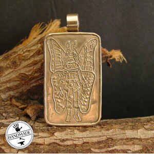 May include: A rectangular bronze-toned metal pendant featuring an embossed design of a winged figure with antennae. The pendant has a smooth, raised border and a cylindrical bail at the top for attachment. The surface has a metallic finish.