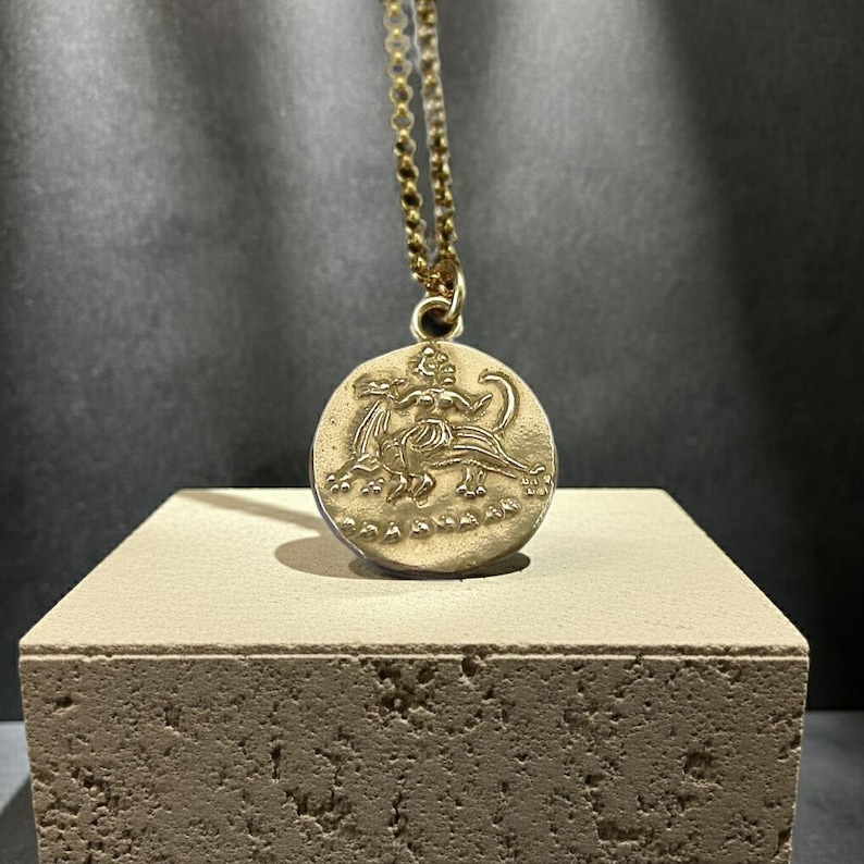 May include: A gold-toned circular pendant necklace featuring a relief design of a mythical creature with a human-like torso and animal body. The pendant hangs from a delicate gold-toned chain, showcasing a textured surface and detailed engraving.