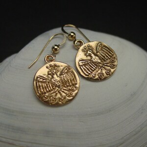 May include: Gold-colored earrings featuring a circular pendant with a detailed phoenix design. The earrings have a hook closure and are displayed on a white, textured surface. The phoenix is depicted with outstretched wings and a crown.