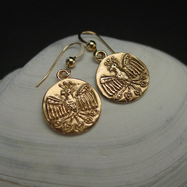 May include: Gold-colored earrings featuring a circular pendant with a detailed phoenix design. The earrings have a hook closure and are displayed on a white, textured surface. The phoenix is depicted with outstretched wings and a crown.