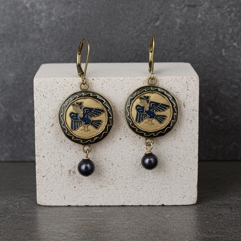 A pair of gold chandelier earrings hanging from gold level back ear wires. The charms are round with a black frame with gold waves surrounding a flying harpy siren, the mythical creature with the body of a bird and the head of a woman. Beneath the charms are navy blue fresh water pearls.
