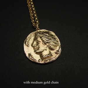 May include: Gold necklace with a large, round pendant featuring a detailed profile of a woman's face. The pendant has a textured, antique-style finish. The necklace is paired with a medium-sized gold chain.