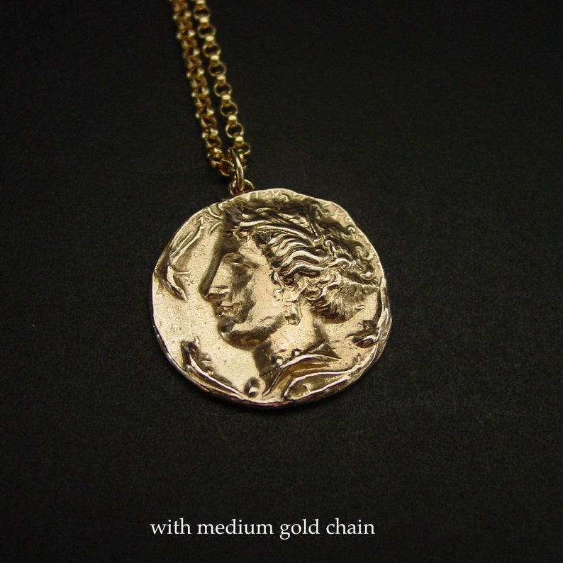 May include: Gold necklace with a large, round pendant featuring a detailed profile of a woman's face. The pendant has a textured, antique-style finish. The necklace is paired with a medium-sized gold chain.