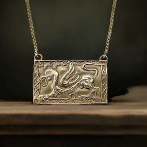 Puede incluir: Un collar con colgante rectangular de color dorado con un relieve detallado de un grifo mítico. El grifo tiene cabeza y alas de pájaro con cuerpo de león. El colgante cuelga de una delicada cadena.