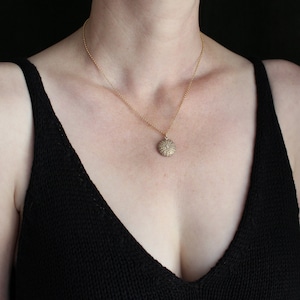 May include: A gold necklace featuring a textured, circular pendant. The pendant is suspended from a fine chain. The person is wearing a black top. The background is dark. The necklace is a piece of jewellery.