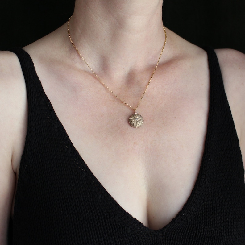 May include: A gold necklace featuring a textured, circular pendant. The pendant is suspended from a fine chain. The person is wearing a black top. The background is dark. The necklace is a piece of jewellery.