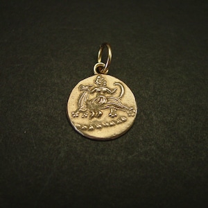 May include: A circular gold-toned metal pendant featuring a relief design of a figure riding a four-legged creature. The pendant has a textured surface and includes a small circular jump ring at the top for attachment to a chain or cord.