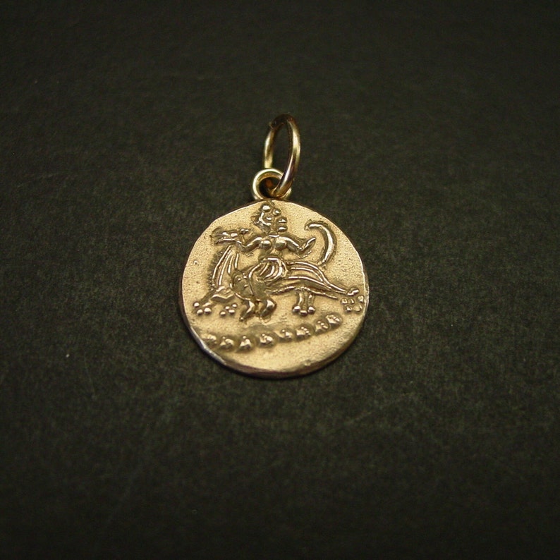 May include: A circular gold-toned metal pendant featuring a relief design of a figure riding a four-legged creature. The pendant has a textured surface and includes a small circular jump ring at the top for attachment to a chain or cord.