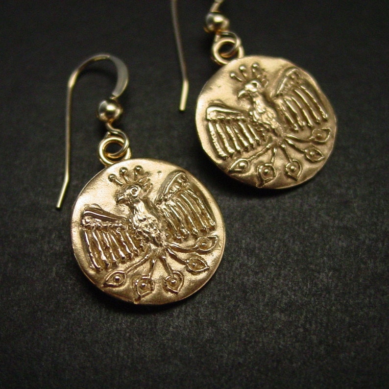 May include: Gold-colored earrings featuring a circular pendant with a detailed phoenix design. The phoenix is depicted with outstretched wings and a crown. The earrings have a simple hook closure and a small gold bead.