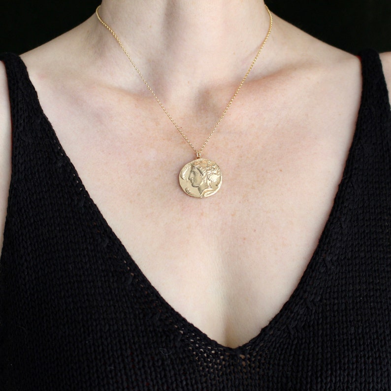 May include: A gold chain necklace with a large, round pendant featuring a profile of a woman's face. The pendant is a detailed, embossed design.