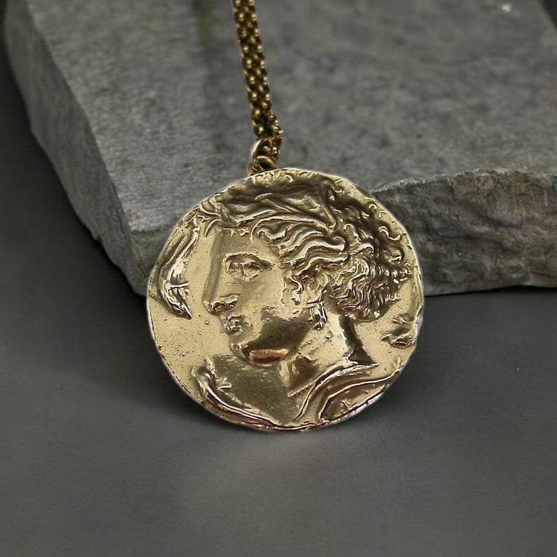 A large gold coin pendant necklace with the raised profile of Persephone wearing dangle earrings. Around her face are three dolphins.