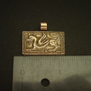 K&ouml;nnte beinhalten: Ein goldfarbener Anh&auml;nger mit einem detaillierten gefl&uuml;gelten Drachen-Design in einem quadratischen Rahmen. Der Anh&auml;nger ist etwa 2,5 cm gro&szlig;.