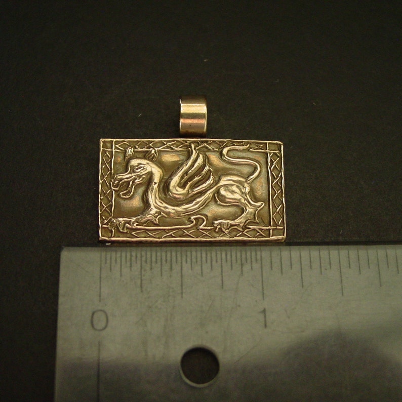 K&ouml;nnte beinhalten: Ein goldfarbener Anh&auml;nger mit einem detaillierten gefl&uuml;gelten Drachen-Design in einem quadratischen Rahmen. Der Anh&auml;nger ist etwa 2,5 cm gro&szlig;.