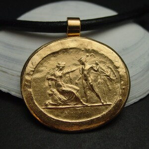 May include: A gold-colored pendant with a detailed relief of two figures in classical attire. The pendant is round and hangs from a black cord. The figures are engaged in a dramatic scene, with one seated and the other reaching out.