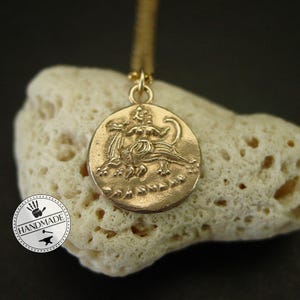 A gold pendant necklace with a round coin-shaped charm featuring a detailed embossed image of a woman riding a mythical creature. The charm is on a white coral-like surface.