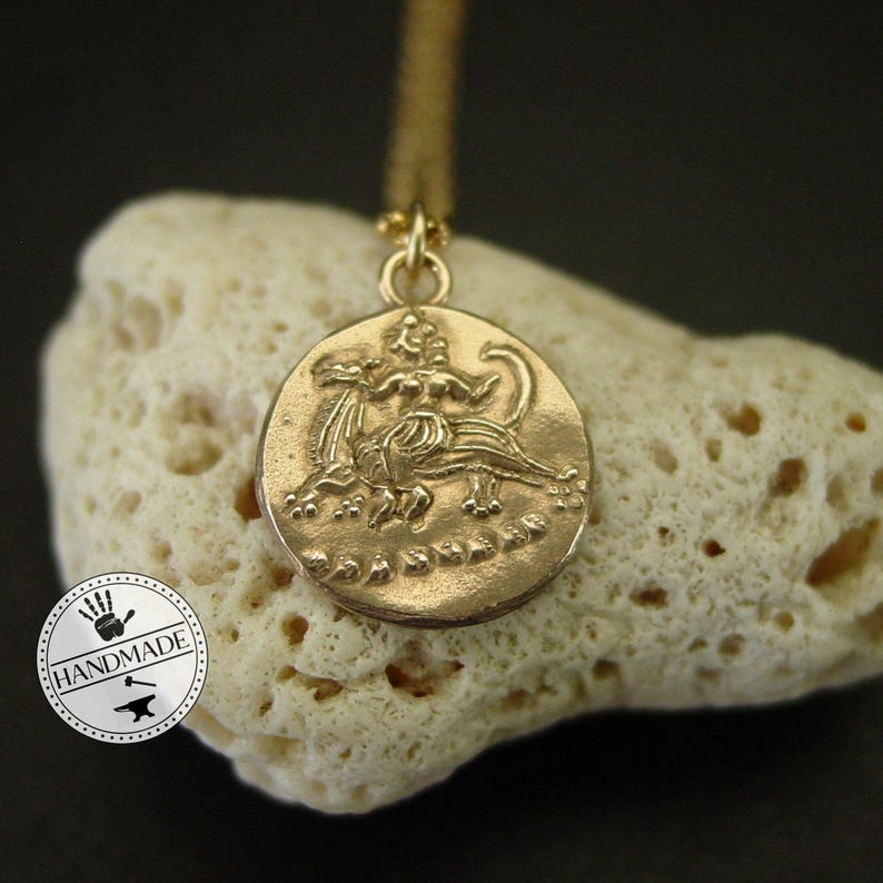 A gold pendant necklace with a round coin-shaped charm featuring a detailed embossed image of a woman riding a mythical creature. The charm is on a white coral-like surface.
