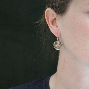May include: Gold-toned coin earrings with a detailed owl design. The earrings are square-shaped with a small hook. The owl is depicted in profile, with intricate detailing. The earrings are worn on a person's ear.