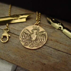 May include: A gold-toned circular pendant featuring a relief of a stylized bird with spread wings and a fanned tail, suspended from a delicate chain. The necklace is positioned alongside two metallic pen nibs on a textured wooden surface.