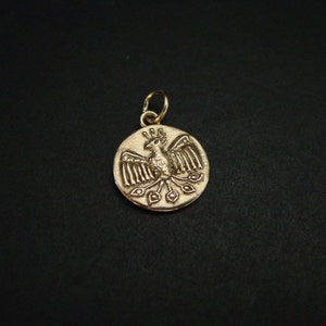 May include: A circular gold-toned metal pendant featuring an embossed design of a stylized bird with outstretched wings, a crested head, and a long tail with three distinct feather-like shapes, attached to a small circular jump ring at the top.
