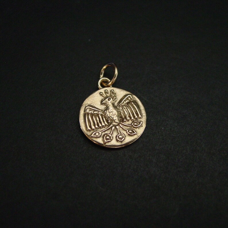May include: A circular gold-toned metal pendant featuring an embossed design of a stylized bird with outstretched wings, a crested head, and a long tail with three distinct feather-like shapes, attached to a small circular jump ring at the top.