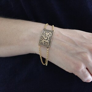 May include: A gold bracelet with a rectangular charm featuring a detailed dragon design.