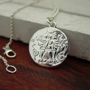 May include: A silver necklace featuring a round pendant with a raised relief design of a multi-armed figure standing between two snakes. The pendant is attached to a delicate silver chain with a lobster claw clasp, showcasing a metallic finish.