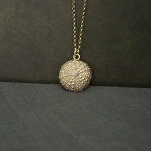 May include: A gold pendant necklace. The pendant is a textured sphere with a small gemstone. The necklace hangs from a fine chain. The background is dark. The pendant is approximately 2.5 cm in diameter.