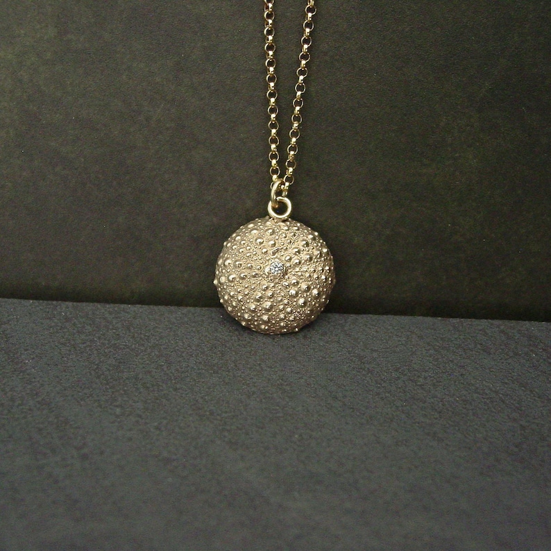 May include: A gold pendant necklace. The pendant is a textured sphere with a small gemstone. The necklace hangs from a fine chain. The background is dark. The pendant is approximately 2.5 cm in diameter.