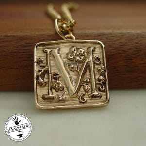 Puede incluir: Un collar con colgante dorado con la letra "M" en un marco cuadrado. La letra está adornada con detalles florales y de vid. El collar tiene una cadena delicada y un logotipo "Handmade".