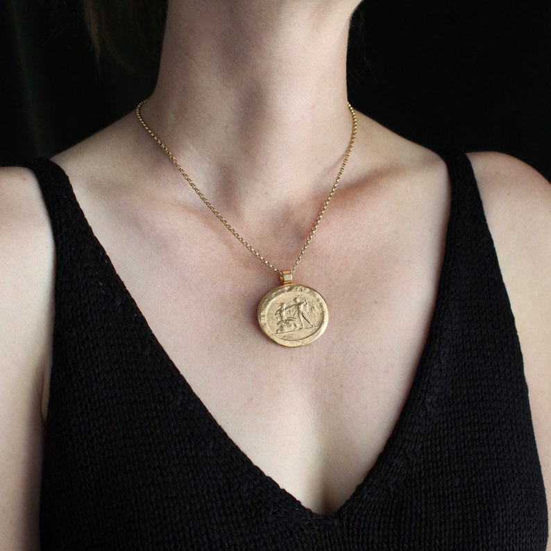 May include: A gold-colored pendant necklace featuring a round, embossed medallion. The medallion depicts a detailed relief scene. The necklace has a delicate chain and is worn against a black top. The overall aesthetic is classic and elegant.