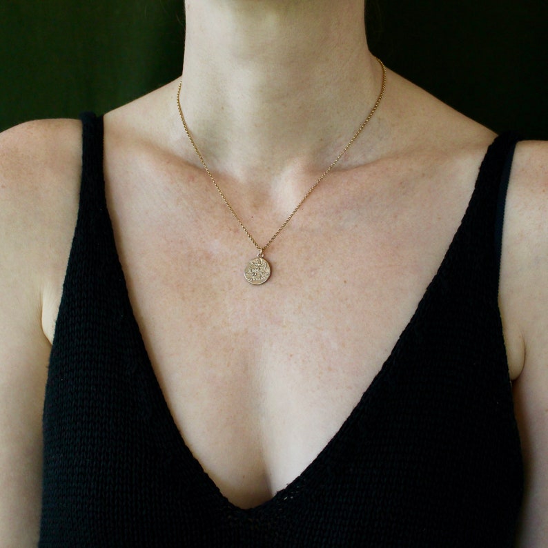 May include: A gold-toned necklace featuring a delicate chain and a small, circular pendant with an embossed design. The pendant has a textured, antique-style finish and hangs centrally from the chain, resting against the collarbone area.