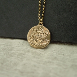 May include: A gold-toned circular pendant necklace featuring an embossed mythological figure riding a creature. The pendant hangs from a delicate gold-toned chain. The design includes a series of small circular beads along the bottom edge of the coin-shaped charm, which has a textured, metallic finish throughout the piece.