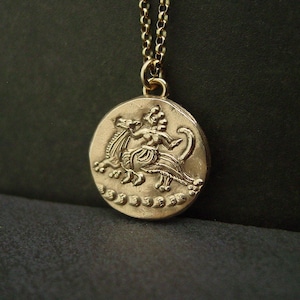 A gold round charm hanging from a thin gold link chain. On the pendant is an embossed figure of a woman riding a flying mystical creature. in the clouds.
