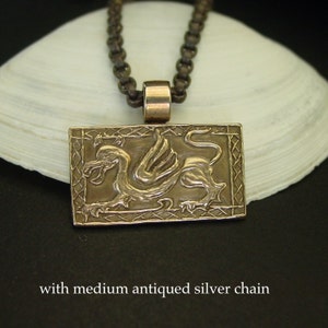 K&ouml;nnte beinhalten: Ein Bronzependant mit einem detaillierten gefl&uuml;gelten Drachen-Design, das in einem quadratischen Rahmen mit einem keltischen Knotenmuster eingefasst ist. Der Anh&auml;nger h&auml;ngt an einer silbernen Kette.