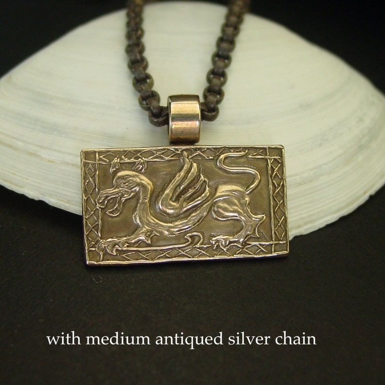 K&ouml;nnte beinhalten: Ein Bronzependant mit einem detaillierten gefl&uuml;gelten Drachen-Design, das in einem quadratischen Rahmen mit einem keltischen Knotenmuster eingefasst ist. Der Anh&auml;nger h&auml;ngt an einer silbernen Kette.