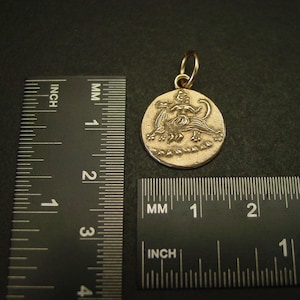 May include: A round gold-toned metal pendant featuring a relief design of a figure riding a mythical creature. The pendant is positioned next to a black ruler showing a diameter of approximately 0.75 inches for the circular charm piece.