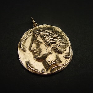 May include: A gold-colored pendant featuring a detailed profile of a woman's face. The pendant is round and has a textured surface.