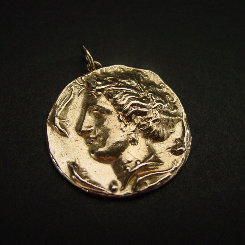May include: A gold-colored pendant featuring a detailed profile of a woman's face. The pendant is round and has a textured surface.