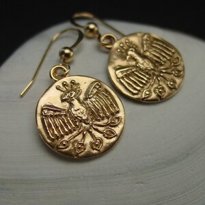 May include: Gold-colored earrings featuring a detailed phoenix design. The earrings have a circular pendant with a bird motif and a hook closure. The phoenix is depicted with outstretched wings and a crown. The earrings are made of a shiny, metallic material.