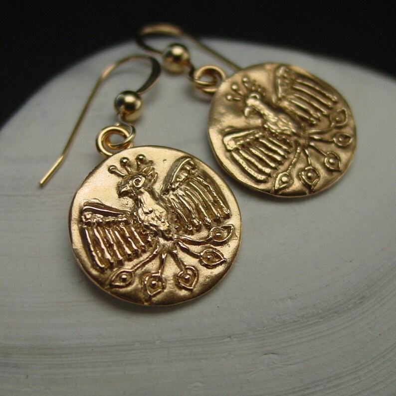 May include: Gold-colored earrings featuring a detailed phoenix design. The earrings have a circular pendant with a bird motif and a hook closure. The phoenix is depicted with outstretched wings and a crown. The earrings are made of a shiny, metallic material.