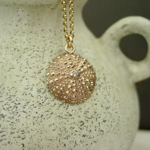 May include: A gold pendant necklace with a textured, round charm, reminiscent of a sea urchin. The charm has a small gemstone in the centre. The necklace chain is also gold, creating a coordinated and elegant design.